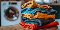 Stack of neatly folded colorful denim jeans and casual pants on wooden surface with washing machine in background, showcasing fresh laundry and home organization.
