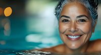 Mature Hispanic woman with silver hair smiling confidently while relaxing in swimming pool with blurred turquoise water and bokeh lights in background. Wellness and active aging concept.
