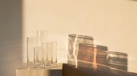 Young woman with light skin tone, professional appearance, arranging clear drinking glasses on neutral surface with warm sunlight casting geometric shadows for interior design and lifestyle content.