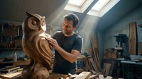 Middle-aged Caucasian craftsman carving detailed wooden owl sculpture in sunlit workshop attic. Artisan woodworker creating handmade bird art piece.
