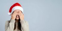 Young woman in Santa hat covering face with hands in playful peek-a-boo gesture against gray background. Holiday stress, surprise, embarrassment concept.