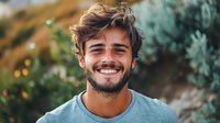 Young Caucasian man with beard and tousled brown hair wearing light blue t-shirt smiles genuinely at camera against blurred natural background in warm evening light.