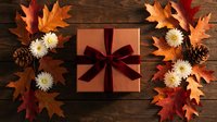 Autumn gift box with burgundy ribbon surrounded by colorful fall leaves, pine cones and chrysanthemums on rustic wooden background for seasonal celebrations.