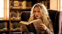 Young Caucasian woman with curly blonde hair sitting in armchair, reading book with shocked expression in cozy library setting.