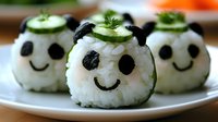 Cute panda-shaped sushi rice balls with nori faces, cucumber slices, and herbs arranged on white plate. Creative Asian food styling for children's menu or party.