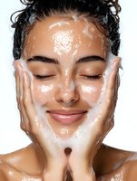 Young Hispanic woman with closed eyes and gentle smile washing face with foam cleanser, demonstrating proper skincare routine on white background.