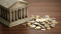 Miniature classical bank building with scattered coins on wooden surface representing financial institutions, savings, investment and economic concepts.