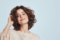Young Caucasian woman with curly brown bob hairstyle and natural makeup smiling confidently against light background, wearing beige top. For beauty and fashion advertising.