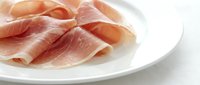 Thinly sliced cured ham arranged on a white plate, showcasing delicate pink meat with a soft texture, perfect for food menus and culinary blogs.