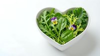 Fresh mixed salad greens with edible flowers in heart-shaped white bowl, overhead view on white background. Healthy eating and nutrition concept.