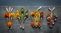 Colorful spices and herbs arranged in wooden spoons on dark slate background creating culinary art display for cooking and seasoning recipes.