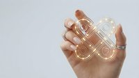 Hand holding transparent glass sculptural object with geometric circular patterns and flowing curves against neutral background.