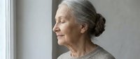 Mature woman with gray hair in bun, looking out window with peaceful expression. Senior female, Caucasian ethnicity, contemplative mood. Suitable for wellness, aging, lifestyle, mental health content.
