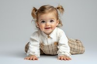 Joyful baby girl with pigtails wearing white sweater and plaid overalls crawling on studio floor, expressing pure happiness against light background.