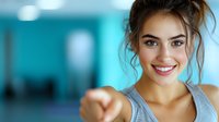 Young mixed race woman with bright smile pointing forward at camera against turquoise background. Casual sporty style with messy bun hairstyle shows confidence and energy.
