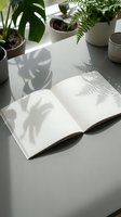 Open blank notebook with plant shadows on white desk surrounded by potted houseplants creating natural light patterns for creative workspace design.