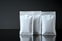 White blank foil or plastic sealed bags standing on reflective black surface against dark background, mockup for product packaging design.
