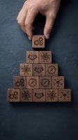 Hand placing wooden block with gear icon atop pyramid stack of engraved business symbols including handshake, compass, and circuit patterns on gray surface for strategy concept.