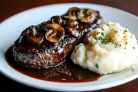 Grilled beef steak topped with sauteed mushrooms in rich brown sauce, served with creamy mashed potatoes garnished with fresh herbs on white plate.