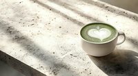 White ceramic cup with matcha green tea latte featuring heart-shaped latte art on concrete surface in natural sunlight with dramatic shadow patterns.