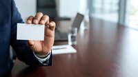 Businessman in dark suit holding blank white business card in office environment with wooden desk and blurred background, suitable for corporate branding and professional networking materials.