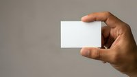 Male hand with medium skin tone holding blank white business card against neutral gray background for branding and corporate identity mockups.