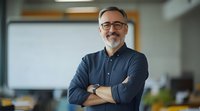 Confident mature businessman with gray beard and glasses wearing casual navy shirt stands with arms crossed in modern office environment. Professional portrait.