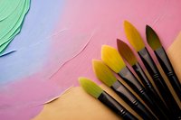 Paint brushes with yellow and green bristles arranged on pink and blue textured background, artistic supplies for creative projects and painting activities.