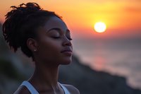 Young African American woman with closed eyes and natural hair in peaceful meditation pose against dramatic orange sunset sky and setting sun over ocean.