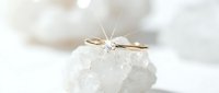 Gold solitaire diamond engagement ring resting on white crystal rock, sparkling gemstone with star light effect on soft white background.