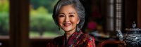 Elegant senior Asian woman with gray hair smiling warmly in traditional red floral dress, sitting in luxurious interior setting for lifestyle and cultural content.