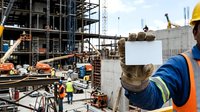 Worker wearing hard hat and safety vest holding blank sign at active construction site with steel framework and multiple laborers in background.
