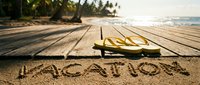 Vacation word written in sand on tropical beach with yellow flip flops on wooden boardwalk, palm trees and ocean in background.