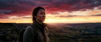Woman with long dark hair and South Asian ethnicity stands on rocky hilltop at dramatic sunset, wearing casual jacket, gazing toward golden and crimson sky. Suitable for travel, adventure, and lifestyle editorial use.