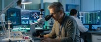Middle-aged Caucasian male scientist examining samples through microscope in modern laboratory with digital displays and analytical equipment.