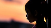 Young Asian girl silhouette against orange sunset sky, side profile view showing contemplative peaceful moment with soft natural backlight creating dramatic mood.