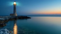 Lighthouse on rocky coast at dusk with glowing beacon reflected in calm turquoise sea water against orange sunset sky and blue twilight horizon.