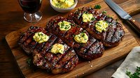 Grilled ribeye steaks topped with melting herb butter on wooden cutting board, garnished with fresh parsley. Served with red wine for fine dining.