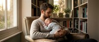 Young father holding newborn baby in cozy home library setting with warm natural light streaming through windows creating intimate bonding moment.