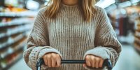 Young Caucasian woman in beige knit sweater holding shopping cart handle in supermarket aisle with blurred store shelves background, cropped view.