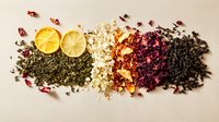 Fresh citrus slices with variety of loose tea leaves and dried flower petals arranged in colorful gradient composition on neutral background.