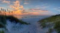 Tranquil beach pathway through sand dunes with sea oats at sunset, footprints leading to ocean waves under dramatic orange and pink clouded sky.