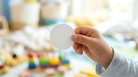 Young child with medium skin tone holding blank white circular card or token, with colorful toys and play items blurred in background, suitable for educational materials and parenting content.