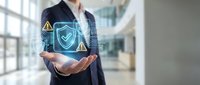 Businessman in dark suit holding glowing shield security icon with warning symbols in modern office environment, representing data protection and cybersecurity solutions.