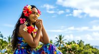 Young woman with long dark hair and medium brown skin tone wearing colorful floral lei and tropical dress, posing outdoors with palm trees and blue sky.