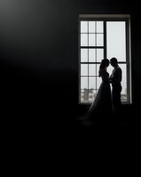 Romantic couple silhouette embracing by large window in dramatic black and white wedding portrait with elegant gown and formal attire.