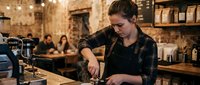 Young female barista in plaid shirt preparing coffee at espresso machine in cozy cafe with warm lighting and customers in background.