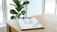 Clipboard with handwritten apology note saying I am sorry for the mistake on wooden desk, pen beside it, green plant and window in background.