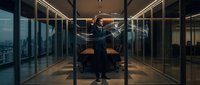 Professional businessman in dark suit standing confidently in modern glass-walled conference room with city skyline view during evening hours.