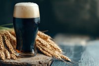 Glass of dark craft beer with thick foam head and wheat ears on rustic wooden surface against dark moody background, selective focus beverage photography.